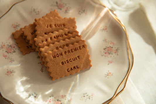 Biscuit en forme de petit beurre avec écrit dessus le message : "mon premier biscuit Carl", posé sur une joli assiette au motif florale et au contour doré.