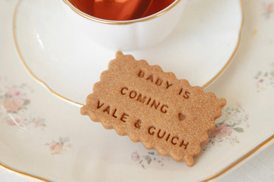 Biscuit en forme de petit beurre avec écrit dessus le message : "baby is coming avec un motif petit coeur et signé avec deux prénoms", posé sur une joli assiette au motif florale et au contour doré avec une tasse de thé dans le fond.