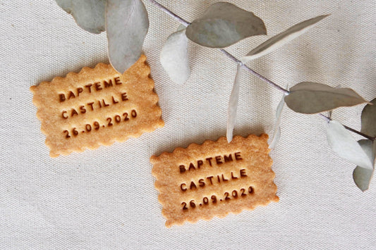 Biscuit personnalisé en forme de petit beurre avec l'inscription "Baptême Castille Septembre", posé sur une table de baptême au style champêtre avec des fleurs séchées.